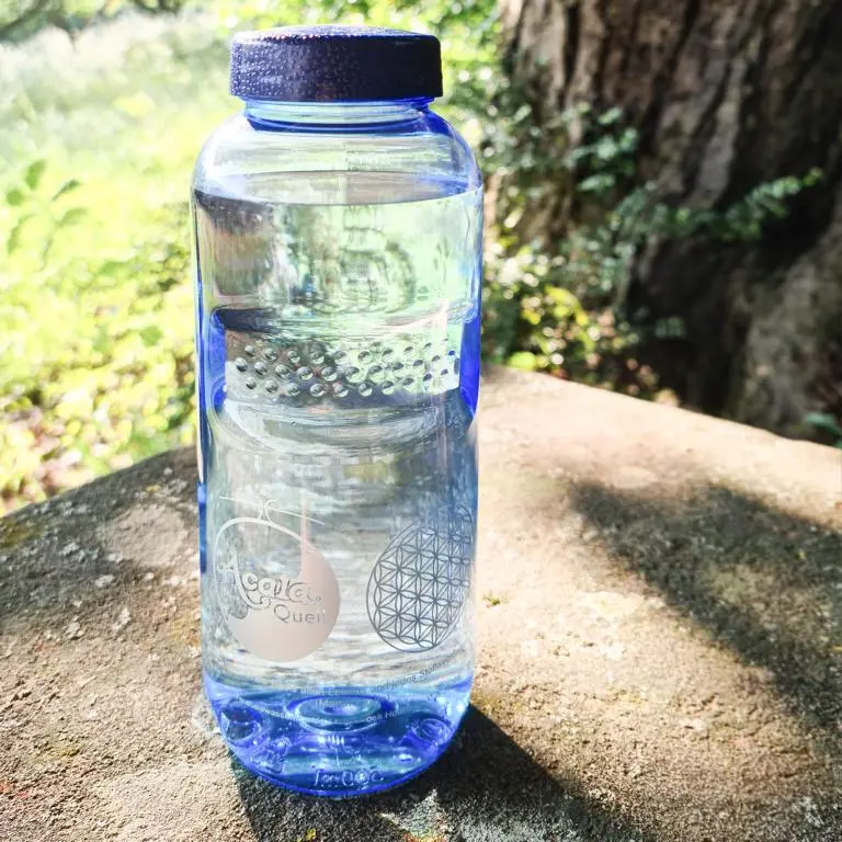 Zu sehen ist eine mit Wasser gefüllte Tritan Trinkflasche in blau mit blauem Schraubdeckel. Sie steht auf einem Stein, im Hintergrund sieht man eine Wiese und einen Baum, die Sonne scheint. Das Acala Logo ist in weiß auf der Flasche zu sehen. 