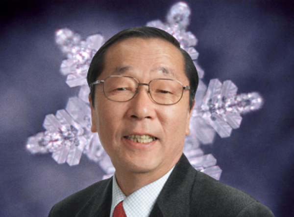 Zu sehen ist Masaru Emoto. Er ist Mitte fünzig, trägt einen dunklen Anzug mit roter Krawatte und eine Brille. Er schaut lächelnd in die Kamera und man sieht einen Wasserkristall vor einem dunklen Hintergrund hinter ihm.