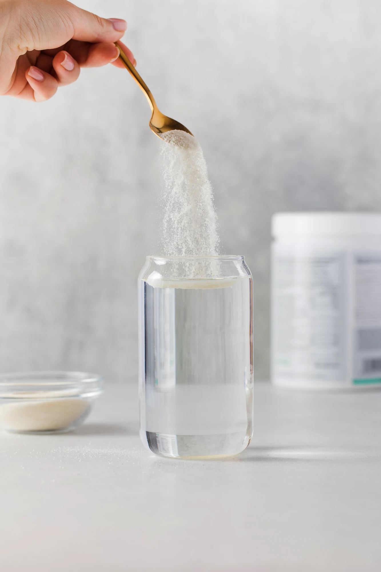 Man sieht eine weibliche Hand, die Pulver von einem goldenen Löffel in ein Glas gefüllt mit Wasser rieseln lässt.