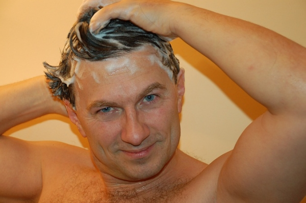 Zu sehen ist ein Mann mit freiem Oberkörper, der sich die Haare wäscht. Er hat kurze braune Haare voller Schaum und schaut lächelnd in die Kamera. Mit den Händen schäumt er die Haare ein.