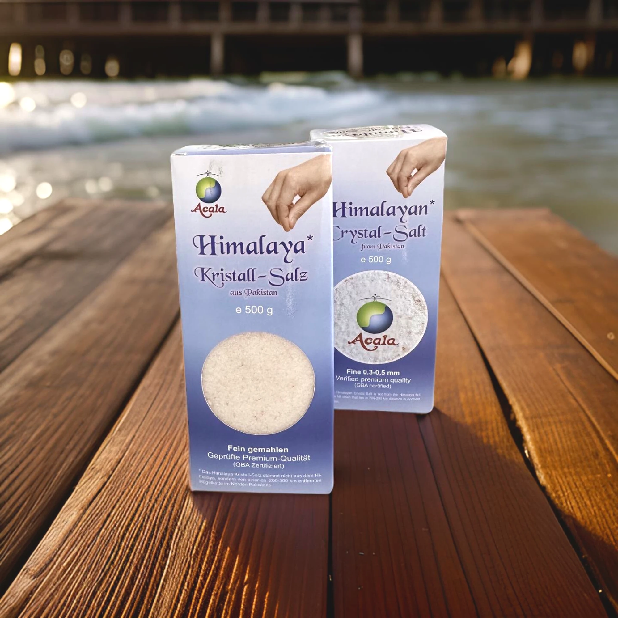 Zu sehen sind 2 Packungen Kristallsalz fein 500g von Acala. Sie stehen auf einem dunklen Holztisch, im Hintergrund sieht man das Meer und eine Seebrücke.
