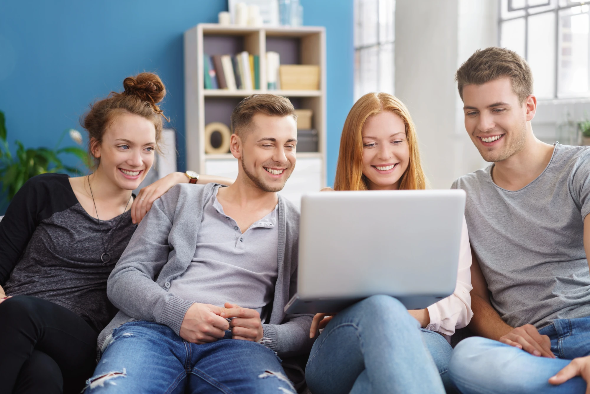 Vier junge Leute in einem Wohnzimmer schauen lächelnd auf einen geöffneten Laptop.