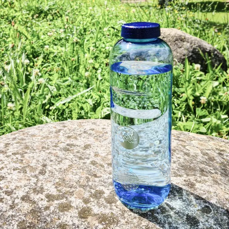 Eine mit Wasser gefüllte Tritan Trinkflasche Grip 1L in blau mit blauem Schraubdeckel auf einem Stein in einer Wiese.