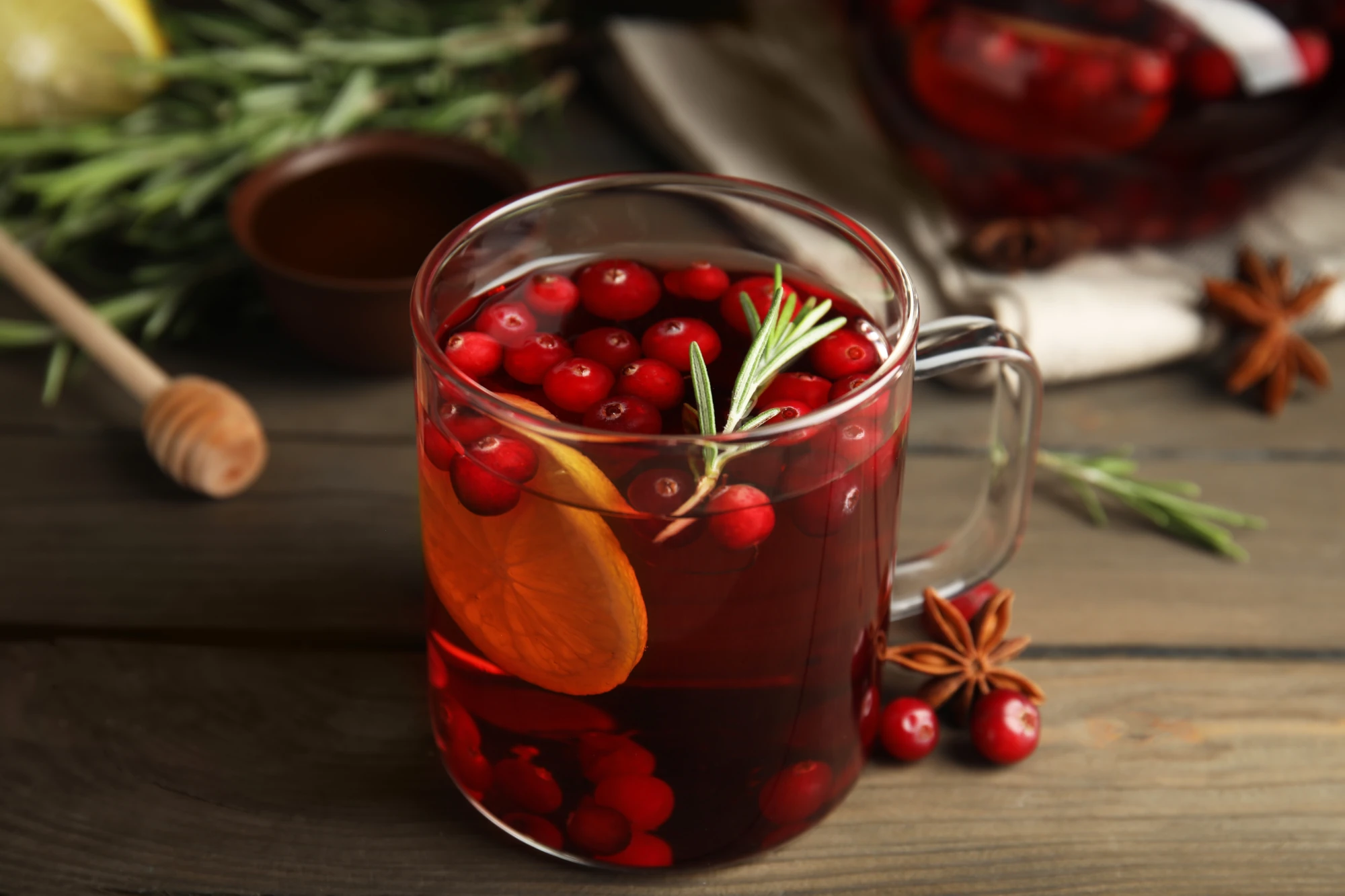 Durchsichtige Tasse mit roter Flüssigkeit, roten Beeren, einer Orangenscheibe und einem grünen Zweig darin auf einem Holztisch. Im Hintergrund weitere Zutaten.
