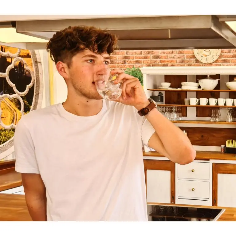 Das Bild zeigt einen jungen Mann um die 20, der in einer Küche steht und aus einem Glas Wasser trinkt. Er hat kurze braune Haare und trägt ein weißes Shirt. Das Bild zeigt das Arlando Trinkglas von Acala.