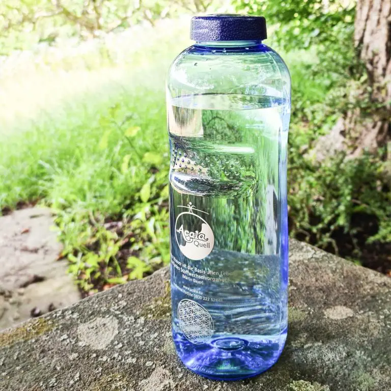 Zu sehen ist eine mit Wasser gefüllte Tritan Trinkflasche in blau mit blauem Schraubdeckel. Sie steht auf einem Stein, im Hintergrund sieht man eine Wiese und einen Baum, die Sonne scheint. Das Acala Logo ist in weiß auf der Flasche zu sehen. 